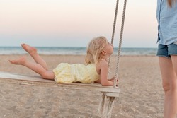 Mother rides her little daughter on swing on the seashore. Cute towheaded girl on rope swing on the beach on sea background