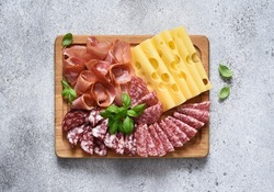 Delicacies. Cheese, prosciutto, salami on a wooden square board on a concrete background.