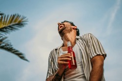 Low angle view of a smiling man holding a cocktail and laughing outdoors against the blue sky	
