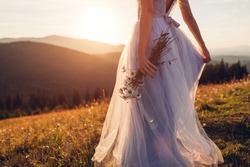 Bride wearing blue wedding dress holding bouquet in mountains at sunset. Woman walking on meadow in flowers