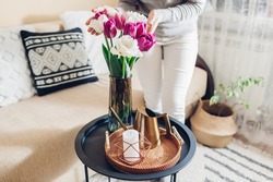 Woman enjoys tulips flowers put in vase on table with tray, watering can and candle. Cozy interior and spring decor at home