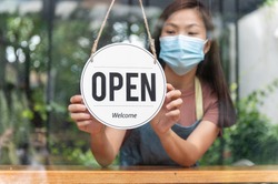Asian small business owner wearing protective face mask and turning sign to open again after the quarantine due to coronavirus pandemic. Woman hanging open sign on the glass window. Focus on sign.