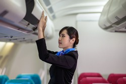 Young beautiful Asian airline cabin crew attendant closing overhead cabinet luggage compartment for checking safety of passenger before flight. Airline business professional uniform occupation concept