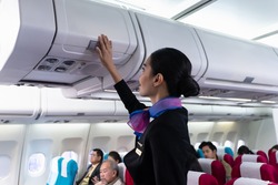 Young beautiful Asian airline cabin crew attendant closing overhead cabinet luggage compartment for checking safety of passenger before flight. Airline business professional uniform occupation concept