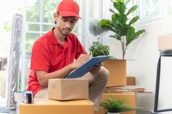 Portrait young Asian man house moving service worker in uniform doing home relocation, checking list cardboard boxes for preparing to move. Package delivery resident relocation moving service concept.