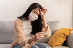 Young Asian woman wearing face mask have a cold and high fever while checking body temperature by using digital thermometer. Daily lifestyle health care concept.