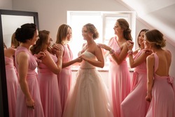 Bride in white wedding dress and bridesmaids in pink dresses posing in hotel or fitting room at wedding day.