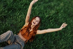 A woman lying in nature on the green grass in the park resting with her hands up and smiling with her mouth open, her red hair flying in the wind in the open air in the sunlight. Freelancer's concept