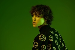 Man fashion and style accessories model with curly hair stylish sweater, hipster teen lifestyle, portrait green background mixed neon light, copy space