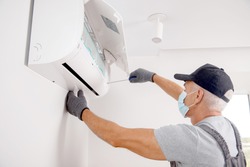 Technician man in medical mask worker repairing and installs air conditioner on white wall.