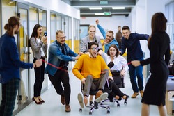 Young colleagues group having fun together, riding on chairs in office, diverse excited office workers enjoying break, laughing, engaged funny activity, celebrating corporate success.