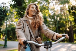 Smiling woman with curly hair in a coat rides a bicycle in a sunny park. Outdoor portrait. Beautiful woman enjoys nature. Lifestyle. Relax, nature concept. 
