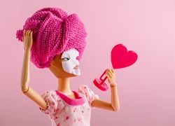 Plastic doll with towel on head and mask on face watching into mirror on a pink background. Spa and beauty care concept.