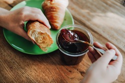 Hands holding a spoon with plum jam and a croissant. Personal perspective view. taking a bite.
