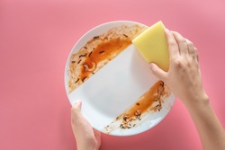 woman using yellow cleaning sponge to clean up and washing food stains and dirt on white dish after eating meal isolated on pink background. cleaning , healthcare and sanitation at home concept