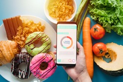 Calories counting and food control concept. woman using Calorie counter application on her smartphone with fresh vegetables, dessert and donuts on dining table