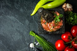 Traditional Latin American mexican Tomato sauce salsa, chilean chancho en piedra in stone mortar and ingredients tomatoes, chile, garlic, onion, sea salt on dark slate stone background
