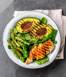 Healthy grilled chicken, grilled avocado and asparagus salad with linen seeds. Balanced lunch in bowl. Gray slate background. Top view.
