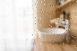 Water drops on wet glass shower door in hotel bathroom with blured bokeh window, sink, faucet and wooden furniture on sunny morning or day. Travel, holiday, vacation, interior design, body care