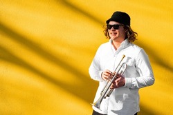 Portrait of young European long-haired cheerful smiling male musician in white shirt, hat and sunglasses holding silver trumpet ready to play standing near yellow wall on street outside on sunny day