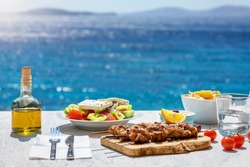 Greek food concept with farmers salad and souvlaki skewers in front of the sparkling, blue Aegean sea during summer time