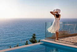 A elegant luxury woman in a white dress enjoys the summer sunset by the pool overlooking the Aegean Sea in Greece