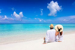 A beautiful tourist couple with hats sits on a tropical beach and enjoys the view to the turquoise ocean during their summer holiday