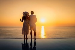 A hugging couple stands by the swimming pool and enjoys the intense sunset over the mediterranean sea in Greece with a glass of wine