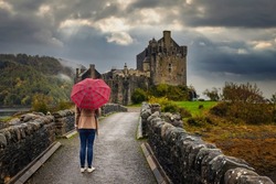 A tourist woman with a scottish pattern umbrella walks towards the Eilean Donan castle on a rainy autumn day, Scotland