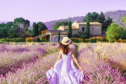 Provence, France. Charming young woman in Blooming Lavender fields at background of beautiful traditional French Provencal house.  Back view of lovely lady wearing waving Boho Chic purple Dress, hat.