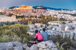 Middle aged couple of travellers from Asia sitting at stone admiring fabulous sunset over ancient Athen's city downtown, view at Parthenon and Agora hill, during twilights. Athens, Greece, Europe.