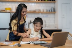 Asian mother and little daughter sit at desk in kitchen studying online together, biracial mom and small girl child handwrite, do homework, learning at home, homeschooling concept