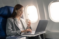 Attractive asian female passenger of airplane sitting in comfortable seat listening music in earphones while working at modern laptop computer with mock up area using wireless connection on board.