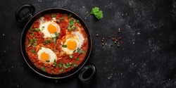 Shakshuka eggs in a pan with toast on a black concrete background. Poached eggs in a spicy tomato pepper sauce. Traditional Jewish scrambled eggs. Top view, flat lay. Banner. Textured object, selectiv