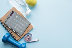Paper notebook, calculator, pen, bottle of water, dunbbell, measuring tape and apple on blue background. Weight loss, fitness, counting calories concept. Top view. Copy space.