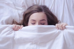 Beautiful young woman lying down in the bed and sleeping. Teen girl with closed eyes covers her face with white blanket in the morning. Do not get enough sleep concept. View from above. Copy space.