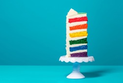 Single slice of birthday cake on a white cakestand minimalist on a blue background. Homemade cake with multicolored layers and buttercream