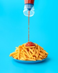Pouring ketchup from a bottle over french fries, minimalist on a blue background. Delicious meal, french fries with ketchup in bright light on a colorful table.