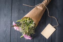 Gifting theme image with a lovely bouquet of flowers wrapped in brown paper and a blank label tied to it, on a black wooden background.