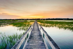 sunset over coastal waters with a very long wooden boardwalk pier in the center during a colorful summer sunset under an expressive sky with reflections in the water and marsh grass in the foreground