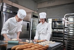 Two professional Asian chefs in white cook uniforms and aprons knead pastry dough and eggs, prepare bread, cookies, and fresh bakery food, baking in an oven in a restaurant stainless steel kitchen.