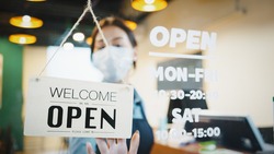 Asian female restaurant owner wear face mask, flip the shop's sign placard from Closed to Open. Business reopen after covid-19 coronavirus pandemic, Covid relief, or new normal lifestyle concept