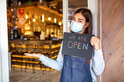 Happy female waitress with protective face mask holding open sign while standing at cafe or restaurant doorway, open again after lock down due to outbreak of coronavirus covid-19