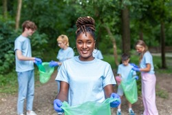 Portrait of beautiful woman with small group of volunteers on background with gloves and garbage bags cleaning up city park - environment preservation and ecology concept. All wearing a blue t-shirts