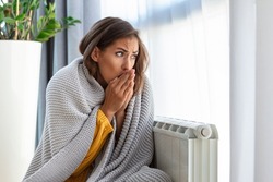 Woman freezing at home, sitting by the cold radiator. Woman with home heating problem feeling cold