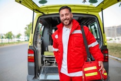 Young man , a paramedic, standing at the rear of an ambulance, by the open doors. He is looking at the camera with a confident expression, smiling, carrying a medical trauma bag on his shoulder.