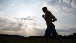 Silhouette of muscular man jogging in the country road at sunset. Profile of male jogger training for marathon run outdoor. Athlete exercising and running against blue sky. Sport and active lifestyle.