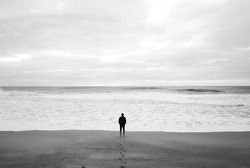 Man goes to the sea on dark sand beach under cloudy sky