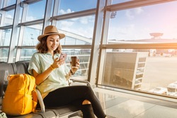 Asian girl waiting for departure at the airport on your vacation. Uses a smartphone and drinks coffee