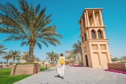 Woman tourist wearing a maroon turban and yellow backpack walks along old wind towers and palms in Bur Dubai and Creek district. Travel and sightseeing spots
